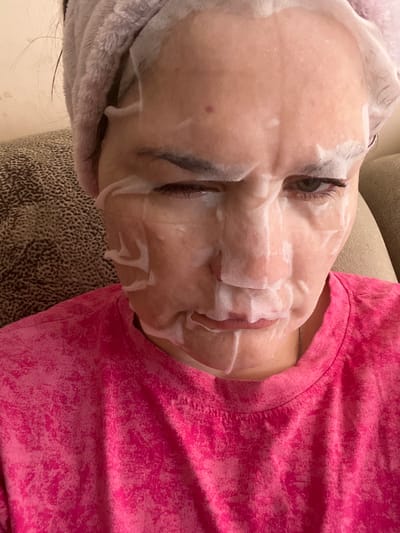 A woman is wearing a pink t-shirt and a light purple towel or head wrap. Her face is covered with a sheet mask, which appears to be foamy in places. She is looking at the camera with her right eye squinted and her mouth slightly open. The background shows a textured brown couch and a light-colored wall. The image was taken indoors. No specific location in Afipskiy, Russia, can be determined from the visual information.