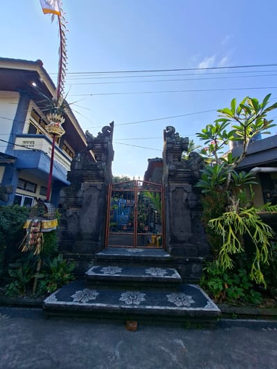 The image shows an ornate entrance gate to a building in Blahbatuh, Indonesia. The gate is made of dark stone with intricate carvings, flanked by two pillars topped with decorative figures. A set of steps with floral patterns leads up to the gate. To the left of the gate, a tall bamboo pole with a flag and offerings is erected, characteristic of Balinese ceremonies. The building behind the gate has a blue and white facade, with a prominent roofline. Lush green vegetation surrounds the entrance, and a frangipani tree with pink blossoms grows to the right of the gate. The sky is clear and blue, with power lines running across it. The scene depicts a typical residential entrance in the region, possibly decorated for a cultural event.
