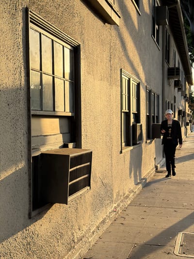 A low-angle photograph depicts a building's facade, possibly in West Hollywood, California, with a man positioned to appear as if he is leaning against the building wall. The building is a light tan color with multiple rectangular windows and air conditioning units placed in some of the window spaces. The man is wearing a black jacket and trousers with a white head covering and appears to be suspended horizontally in the frame. A shadow of the man is cast on the building. To the right, a grey concrete wall is visible, marking a potential corner of the building. The lighting suggests late afternoon, casting elongated shadows across the textured surface of the building.