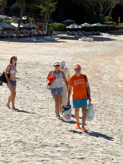 Four people are on a sandy beach near a row of lounge chairs and umbrellas. The individuals appear to be a mix of adults, with two men and two women.
The first man from the top is wearing an orange t-shirt with the text "KULT MADE IN POLAND" and blue shorts. He is holding a white bag and wearing orange sneakers. Next to him is a woman in a white top with a patterned design and denim shorts, also wearing a blue cap. She is holding a red towel and a small white bag.
Below them, a woman is lying on the sand wearing a white polka dot bikini and denim shorts. She is wearing sunglasses and a black bag is next to her. The fourth person's lower body is visible in the upper left quadrant of the image, wearing a light-colored garment and resting on the sand.
The scene is set on a beach with sand, vegetation, and lounge chairs. The lighting suggests it is daytime, likely late afternoon due to the long shadows cast on the sand. The environment appears to be tropical or subtropical, possibly in Baan Plai Laem, Thailand, as indicated by the palm trees and lush greenery in the background. The people seem relaxed and are enjoying their time at the beach.