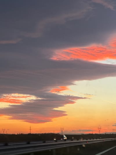The image shows a sunset view captured from inside a moving vehicle, likely on a road near Malenovo, Bulgaria. The sky is predominantly orange and yellow, with streaks of orange-lit clouds against a gray cloud mass. The road infrastructure, including the guardrail and possibly a lane marker, is visible on the right side. Utility poles and overhead power lines are silhouetted against the sunset in the distance. A vehicle with headlights on is visible ahead on the road. The landscape includes trees. The overall scene suggests twilight or dusk.