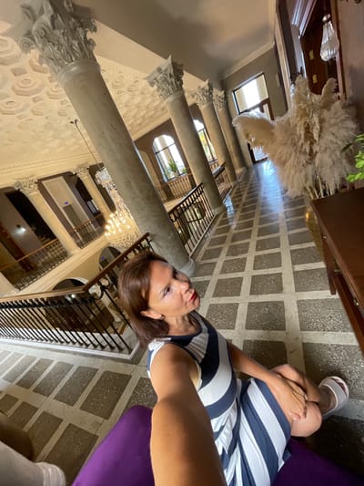 The image shows a woman sitting on a purple cushion in a hallway with a patterned tiled floor and decorative columns. She is wearing a striped dress and white sandals. In the background, there is a chandelier, a staircase with railings, and windows. To the right, there is a wooden table with dried plants. The scene appears to be set in a hotel or a similar building with ornate architecture. The location is Gagra, Abkhazia.