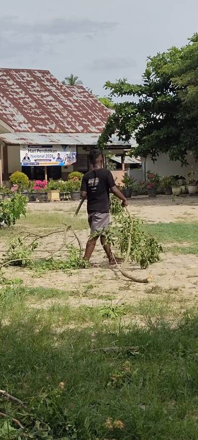 A person is clearing fallen branches from the ground in an outdoor setting. The individual, wearing a black t-shirt and grey shorts, is carrying a large branch with green leaves. They are walking on a dirt or sand-covered ground with patches of grass. In the background, there is a building with a rust-colored, corrugated metal roof. A banner is visible on the building, displaying text in Indonesian, including "Hari Pendidikan Nasional 2026" which translates to "National Education Day 2026." The building appears to be some sort of school or public institution, with potted plants and flowers in front of it. Lush green trees are also present, with one large tree casting a shadow. The sky is overcast with a few clouds, suggesting it might be a warm day. The scene appears to be in a tropical or sub-tropical environment, possibly Langnibong, Indonesia, as indicated by the location context. The activity suggests grounds maintenance or cleanup.