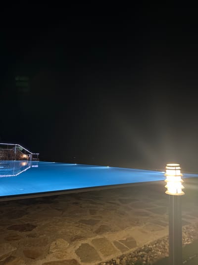 The image depicts a night scene with a large, rectangular illuminated object that appears to be a swimming pool or a similar reflective surface. The water is a vibrant blue, and reflections of lights and structures are visible on its surface. To the right of the pool, there's a stone wall with rough-hewn rocks of various sizes. A modern-style outdoor lamp with multiple horizontal glowing elements is mounted on a pole above the wall, casting a warm light. The background is predominantly dark, suggesting nighttime, and there's a hazy quality in the air, possibly indicating fog or mist. In the lower part of the image, a building with stone cladding and illuminated windows is visible, and its reflection is mirrored in the water. The perspective is from a slightly elevated viewpoint, looking down and across the scene. There is no visible text in the image. The overall mood is serene and possibly tranquil due to the nighttime setting and the gentle illumination.