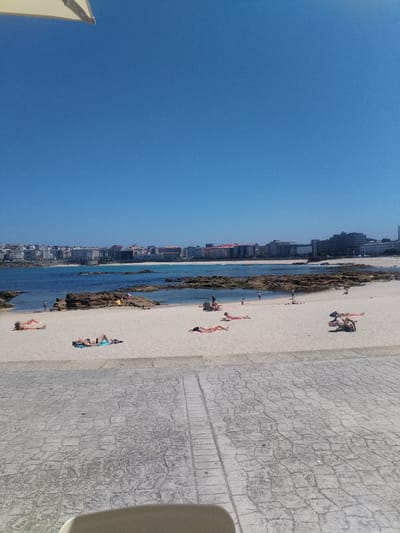 This media file depicts a sunny day at a beach, likely in A Coruña, Spain, given the context. The scene is set against a backdrop of a clear blue sky and a distant cityscape with numerous buildings. The foreground features a paved walkway with a textured, stone-like pattern. Beyond the walkway is a sandy beach populated by several individuals.
The subjects are primarily people sunbathing on the sand. Some are lying on towels, while others are directly on the sand. A few individuals appear to be wading in the shallow, calm water near a rocky shoreline. The overall activity is leisure and relaxation, with people enjoying the beach and warm weather.
Notable details include the bright sunlight, indicating a clear and warm day, likely midday or early afternoon. The water appears inviting and the sand is a light, pale color. The presence of a beach umbrella in the upper left corner suggests amenities for sun protection. The cityscape in the distance provides a sense of urban proximity to this natural setting. The mood is peaceful and serene, reflecting a typical vacation or day-off scene. There is no visible text within the image.