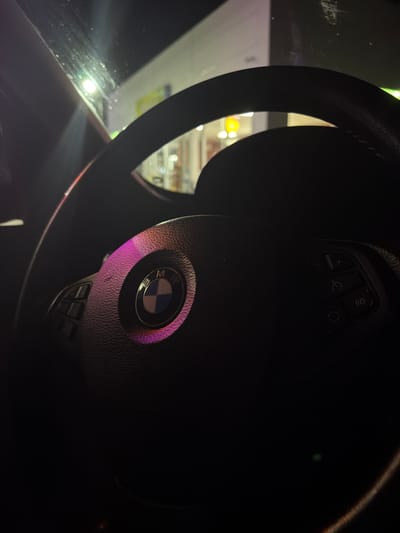This is a close-up, interior shot of a BMW steering wheel, taken at night. A purple light illuminates the BMW logo on the steering wheel and parts of the surrounding dashboard. The exterior environment, visible through the windshield, appears to be an urban setting with bright lights, suggesting the car is parked or moving at night in a well-lit area. The overall lighting is dim, with the exception of the bright exterior lights and the purple illumination on the steering wheel. There is no visible person in the image.