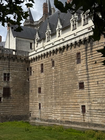 This image shows a view of the Château des Ducs de Bretagne in Nantes, France. The scene captures a section of the castle's imposing stone walls, characterized by their rough-hewn blocks and horizontal banding. Several small windows with iron bars are visible, suggesting a historical defensive or prison function. The castle features elaborate architectural details along its upper sections, including crenellations and decorative molding. In the foreground, green grass and a narrow moat or channel of water are present, hinting at the castle's fortifications. The sky above is overcast, contributing to the somber and historic atmosphere.