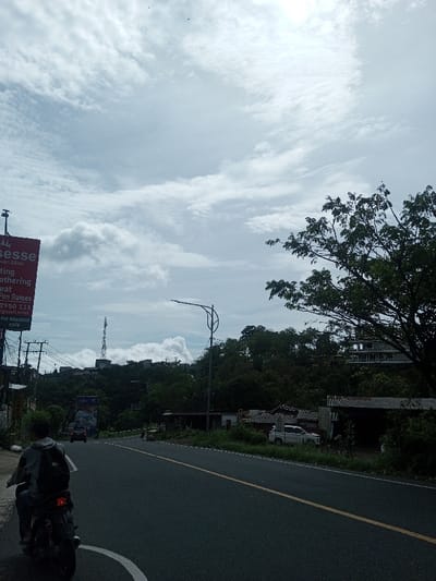 This image depicts a street scene in Sandamal Eliya, Sri Lanka, captured on an overcast day.
In the immediate foreground, on the left, a large red billboard is partially visible, displaying "ESSE Resort Elioto," an address, "Eating," "Gathering," "Fun Games," a phone number "2950 111," an email, and "THE WEEKEND CAFE." Adjacent to the billboard is a utility pole with multiple overhead wires. A person, viewed from behind, wearing a grey hoodie and a black backpack, is seated on a dark motorcycle at the far left, facing away from the camera, positioned on the road's left shoulder marked by a solid white line. The motorcycle's red taillight is illuminated.
The asphalt road, marked with a solid yellow center line, extends into the midground, curving uphill to the right. A dark blue or black vehicle is observed traveling away from the camera further up the road, followed by another motorcycle. A pedestrian is visible on the right shoulder further down the road.
To the right of the road, dense green vegetation covers a hillside. Several small structures with corrugated roofs are nestled among the trees. A white SUV is parked facing the camera, partially obscured by one of these structures. A tall, black street lamp with a curved arm extends over the road from the right side.
In the background, a prominent communication tower stands atop a distant, tree-covered hill, slightly to the left of the frame's center. Further right, through the foliage, the skeletal structure of a multi-story building, possibly under construction, is visible on another hill. The sky is largely covered with light grey, stratocumulus clouds, with some brighter areas indicating diffused sunlight. Two small birds are discernible high in the sky.