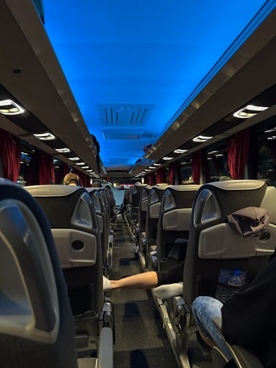 The image shows the interior of a bus, likely traveling through Edirne Merkez, Turkey. The perspective is from a seat looking down the aisle, with rows of empty and occupied seats visible. The ceiling is illuminated with blue light, and red curtains line the windows. Passengers are seated, some with their legs extended. A water bottle is visible in a seat pocket. The overall atmosphere suggests a night or evening journey.