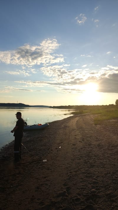 The scene depicts a late afternoon or early evening on a sandy and gravelly riverbank in Шолья, Russia. The sun, low on the horizon, radiates intense light from the right side of the frame, casting a warm glow and creating lens flare. The sky above is a clear blue, adorned with scattered white and grey cumulus clouds.
In the foreground, the ground is dark sand and pebbles, exhibiting footprints and minor debris, including a small, clear plastic container. One individual, appearing male, stands in partial silhouette on the left side of the shore, dressed in dark attire, including a t-shirt, trousers with a reflective strip near the ankle, and a baseball cap. He is holding a fishing rod or similar long object, facing towards the water but looking slightly over his left shoulder towards the camera.
Adjacent to the person, a grey inflatable boat with an attached outboard motor is partially beached on the sand. Another fishing rod lies on the ground near the individual's feet. The calm water of the river or large lake reflects the sky and the warm sunlight, with sparse reeds and vegetation visible along the immediate waterline.
The opposite bank of the water body features a distant, undulating landscape covered with trees and foliage, and what appears to be a power line tower is faintly visible against the horizon. The right bank extends into a grassy area transitioning into denser trees in the distance. The overall environment suggests a tranquil outdoor setting, likely for fishing or recreational activity.