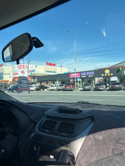 The image, taken from inside a car, shows a street in Аксукент, Kazakhstan. Several stores are visible, including a large "Sulpak" electronics store with red signage, "Media Mart," and a flower shop called "TIM ROSE." Traffic is moderate, with cars lining the street. A rearview mirror and dashboard are prominent in the foreground of the image. An air freshener hangs from the rearview mirror. There is a phone number written behind the dashboard, "87024908958." The sky is clear and blue.