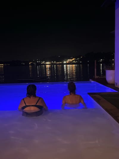 Two women are in a swimming pool at night, facing away from the camera. The pool is illuminated with blue light. In the background, there is a body of water with lights reflecting on its surface, and a distant shoreline with more lights, suggesting a town or village. The overall setting appears to be a tropical location, potentially in Nusa Penida, Indonesia, given the presence of water and lush vegetation visible on the distant hills. The women are submerged up to their shoulders in the pool, and their swimsuits are visible. One woman is wearing a black bikini top, and the other is wearing a yellow bikini top. The atmosphere is tranquil and relaxed, with the dark sky and illuminated water creating a serene ambiance.