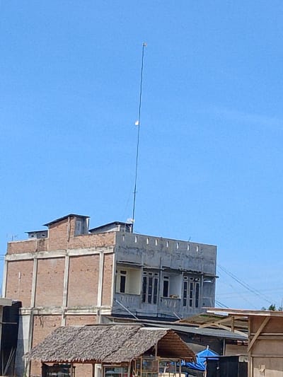 A tall, thin antenna extends vertically into a clear blue sky, topped with a small, light-colored object. The antenna is positioned behind a multi-story building, which appears to be constructed with exposed brick on the left side and a gray concrete facade on the right. The building has multiple windows, many of which are dark. In the foreground, a thatched-roof structure, possibly a small stall or hut, is visible, with other buildings and structures partially obscured behind it. The scene is set against a backdrop of a bright, sunny day, with power lines visible in the distance to the right.