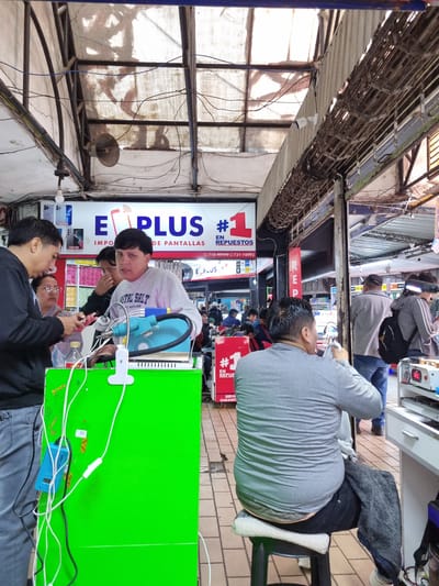 The image depicts a bustling street scene in Santa Cruz de la Sierra, Bolivia, specifically within an area characterized by numerous small shops and stalls. The foreground is dominated by a bright green counter or display surface, cluttered with white electrical cords and a small blue electronic device with a digital display.

Several individuals are present. On the left, a man in a black hooded sweatshirt is looking down at a mobile phone he is holding. Beside him, a woman wearing glasses is also observing the phone. In the center, a young man with dark hair and a light gray long-sleeved shirt is looking towards the camera with a serious expression. He has his hand partially covering his mouth. Behind him, another person appears to be interacting with a mobile phone.

A prominent sign overhead reads "E PLUS" in large blue letters, accompanied by a stylized red graphic of a phone and the number "1" in a red square with the text "#1 EN REPUESTOS". Another smaller sign displays a phone number, "710-99999 731-76992".

Further in the background, other individuals are visible, some standing and others seated. A man in a gray sweatshirt is seated on a white stool, facing away from the camera, holding a white object. Another person with a backpack is visible in the distance. The environment appears to be an open-air market or a covered walkway with exposed electrical wiring and corrugated metal roofing. The ground is tiled. The overall impression is one of a commercial area focused on electronics repair or sales.