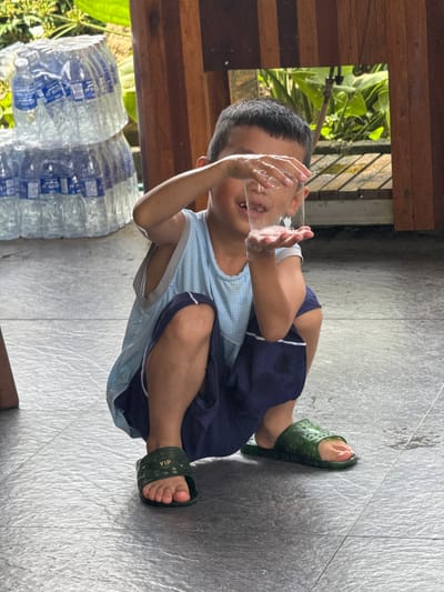 A young child, likely a boy, is depicted sitting on a tiled floor in what appears to be an outdoor or semi-outdoor setting in Du Già Commune, Vietnam. He is wearing a light blue mesh tank top, dark blue shorts with white trim, and green rubber slide sandals.

The child is actively engaged in playing with water, holding a clear glass with both hands. Water droplets and bubbles are visible on his hands and the glass, and his face is partially obscured by the glass as if he just took a sip or is splashing water. The tiled floor around him shows wet patches, indicating spilled water.

In the background, to the left, several large plastic water bottles are stacked, with labels clearly showing "WATER." Behind them and extending into the upper left background, lush green foliage and plants are visible, suggesting a natural outdoor environment. Wooden structures, possibly beams or parts of a building's support, are also present, framing the green background. The bright, even lighting indicates it is daytime.

Notable visible text includes "VIP" embossed on the strap of the child's green sandals and "WATER" on the labels of the stacked plastic bottles. The scene conveys a moment of playful interaction with water on a warm day.