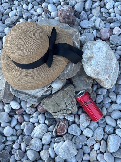 A straw hat with a black ribbon and a red can of Coca-Cola Zero Sugar are placed on a bed of smooth, grey and white pebbles on a beach in Perazića Do, Montenegro. The hat is positioned on top of several larger, grey rocks, with its brim casting a shadow. The Coca-Cola can is leaning against a large, white, rough-textured rock. The pebbles vary in size and shape, with some pinkish-grey stones interspersed among the predominantly grey ones. The scene is captured from a high-angle, overhead perspective.