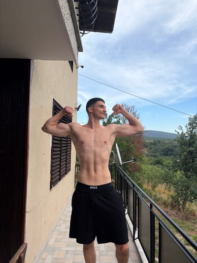 The image shows a young man with short dark hair and a light complexion, shirtless and wearing black shorts with "BOSS" written on the waistband. He is flexing his biceps, looking up and to his left with a slight smile. The background suggests an outdoor setting with a building to the left, a balcony with a railing, and a wooded hillside with trees under a partially cloudy sky. The time of day appears to be daytime, likely afternoon, given the lighting. The scene is set on a tiled patio.