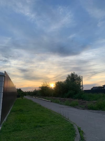 This is a vertical, eye-level, medium shot image of a paved path with a metal fence on the left and trees and vegetation on the right. The sun is setting in the background, casting a warm glow on the sky, which is filled with clouds. The paved path is bordered by a strip of green grass on the left and a disturbed area of dirt with red and white caution tape on the right. Several large pipes lie parallel to the path in the disturbed area. The metal fence on the left is dark brown and appears to be made of vertical slats. The trees and vegetation on the right are lush and green. In the distance, several houses with dark roofs can be seen. The overall scene suggests an outdoor setting in a rural or suburban area, possibly during construction or landscaping work. The time of day is sunset.