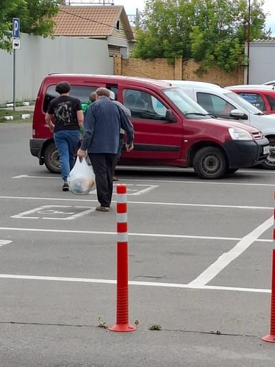 This image captures a scene in a parking lot, likely in Pavlovsky Posad, Russia, given the context. Three individuals are visible, two of whom are actively engaged in an activity. One person, wearing a dark blue jacket and black pants, is bent over, appearing to interact with a large, clear plastic bag. Another person, dressed in a black t-shirt with a graphic design and blue jeans, is also bent over, supporting the plastic bag from below. A third individual, wearing a green shirt, is partially visible behind the person in the blue jacket.

The plastic bag is a prominent object, appearing to contain various items and possibly food. The individuals seem to be loading or unloading this bag from the open rear passenger door of a red van or car. The van itself is a significant element of the scene, its red paint contrasting with the gray asphalt of the parking lot. White lines marking parking spaces are clearly visible on the ground, indicating a designated parking area.

In the background, a building with a light-colored wall and a reddish-brown tiled roof is situated to the left. Trees with lush green foliage are also present, suggesting it is a temperate season, likely spring or summer. Red and white striped traffic cones are placed along the edge of the parking spaces, acting as barriers or markers.

The lighting suggests it is daytime, with no strong shadows indicating the time of day. The sky is not clearly visible, so weather conditions are difficult to ascertain, but the general appearance of the scene is bright enough for clear visibility. The expressions of the individuals are not discernible, making it impossible to infer their emotions. No visible text is present on the people or the objects within the frame, apart from a faint logo on the side of the van which is too blurred to read. The scene appears to be one of everyday activity, possibly involving grocery shopping or moving items.