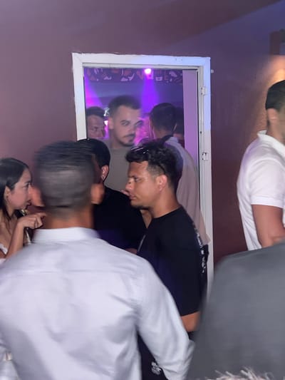 The image depicts a crowded indoor scene, possibly a nightclub or bar, with people gathered in close proximity. The lighting is dim with purple and pink hues, suggesting a nightlife setting. Through an open doorway, a dimly lit room with what appears to be a stage or dance floor with flashing lights is visible. In the foreground, a person in a white pinstripe shirt is facing away from the camera, obscuring the view of those behind them. To the left, a woman in a white top is speaking with someone. To the right, a man in a black t-shirt is looking towards the right, and partially visible is a man in a white polo shirt. The overall impression is of a lively social gathering. The location is identified as Herceg Novi, Montenegro.