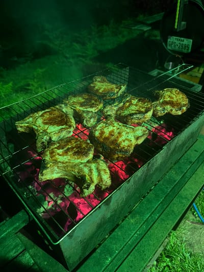A close-up, high-angle shot shows several pork chops grilling over glowing red coals on a dark metal grill. The meat is a golden brown with visible grill marks and appears to be cooked to perfection. The grill is situated on a rustic wooden surface, possibly part of an outdoor seating area. To the left of the grill, a dark, round object is visible, attached to which is a green Texas license plate with white lettering that reads "TEXAS GPG-2985". The surrounding environment is dimly lit, suggesting it is nighttime or dusk. The overall mood is one of casual outdoor dining, likely a barbecue or cookout. The presence of the Texas license plate, despite the location context provided as Ortabatumi, Georgia, is a notable detail. The lighting casts a green hue over the scene, obscuring finer details of the background which includes hints of grass and possibly a blue hose. The focus remains squarely on the appetizing grilled pork chops.