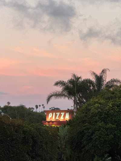 The image depicts a sign that reads "PIZZA" in bright yellow neon letters, illuminated against a warm orange background. The sign is positioned above lush green foliage and palm trees. The sky above is a soft gradient of pink and gray clouds, indicative of a sunset or sunrise. The scene appears to be in Encinitas, United States of America, a coastal city in Southern California known for its relaxed atmosphere and often picturesque skies. The overall impression is one of a casual, possibly commercial, establishment set against a natural, tranquil backdrop during twilight hours.