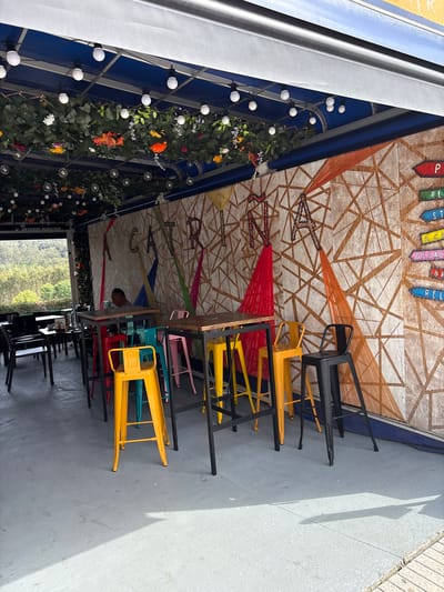 The image shows the outdoor area of a restaurant in O Pino, Spain. Several tables with stacked, colorful chairs are present on a concrete patio. The ceiling is decorated with an intricate design featuring painted plywood and string art in various colors. Vines and light bulbs adorn the walls, creating a decorative ambiance. A view of a green, forested area is visible through the restaurant's entrance. A person is partially visible between the tables and chairs.