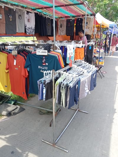 A street vendor is selling clothing on a metal rack on a sidewalk in Kendal, Indonesia, located at the coordinates -6.935 latitude, 110.203 longitude. The vendor is displaying a variety of items, including shirts, shorts, and underwear. The clothing is hanging on hangers, with a price tag of 25,000 attached to a teal shirt with the word "UNWRECKED" printed on it. A price tag of 3 for 60,000 is displayed on the rack with a variety of shorts in various colors, with an additional price tag of 15,000 nearby. A person appears to be looking at the clothing, standing in front of the rack, to the right. The sidewalk is gray concrete, and there is a folding chair nearby. There is a green bag on the ground in front of the rack and a second, darker bag behind it. The vendor is operating under a green and red awning, with the roof visible. There are several other stalls to the right, behind the vendor.