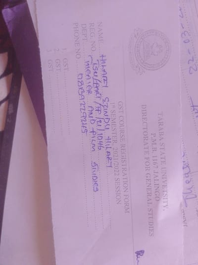 The image shows a close-up of a document with handwritten entries. The document appears to be a GST Course Registration Form for the 1st Semester of the 2021/2022 session at Taraba State University, P.M.B. 1167, Jalingo, Directorate for General Studies.

The form has fields for Name, Reg. No., Dept., and Phone No. The name "HILARY EZINDU HILARY" is written in blue ink. The registration number is "TSU/PART/TP/21/1046". The department is listed as "THEATER AND FILM STUDIES". The phone number is "98139279245". There are also numbered entries for GST courses: 1. GST, 2. GST, and 3. GST.

The document is light purple and shows some creases and smudges, indicating it is a used paper form. There is a circular emblem on the right side of the document, possibly a university seal. Faintly visible text in purple ink on the right side reads "22-03-22" and "TARABA STATE UNIVETSITY, P.M.B. 1167 JALINGO DIRECTORATE FOR GENERAL STUDIES GST COURSE REGISTRATION FORM 1st SEMESTER, 2021/2022 SESSION".

The overall context suggests an administrative or academic setting within Nigeria, specifically Taraba State University. The information captured is factual data related to student registration.