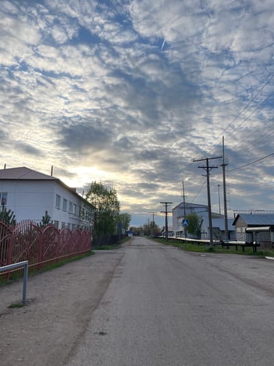 The image shows a street scene in Ulukulevo, Russia. The street runs straight into the distance, with a dirt road on the right side and an open sky dominating the left side. A white building with a red roof and a decorative red metal fence stands along the street on the lower part of the image. Utility poles and wires line the left side of the road, and other buildings are visible further down the street. The sky is filled with scattered, puffy clouds, with sunlight filtering through them, creating a bright glow.