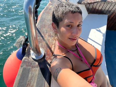 A close-up shot features a person, likely female, with short, dark, closely-cropped hair, looking directly at the camera with a slight, relaxed smile. Her complexion appears tanned, and her eyes are dark. She is wearing an orange bikini top with black trim, and a pink lanyard with a white or light-colored clip hangs around her neck. Two necklaces are visible: a thin silver-colored chain and a dark, possibly beaded, necklace. A tattoo is partially visible on her right upper arm.
She appears to be on a boat or watercraft. To her left, sparkling blue-green water stretches out, reflecting bright sunlight in numerous shimmering points. A red and blue marine buoy floats in the water next to the boat, secured by a thick, dark rope that is tied to a shiny silver metal stanchion. The stanchion rises from a weathered wooden deck, which shows distinct grain and possibly some non-slip strips. To her right and in the background, parts of the boat's structure are visible, including white painted sections and more wooden elements, with strong shadows indicating bright overhead sunlight. The overall impression is of a sunny day out on the water.