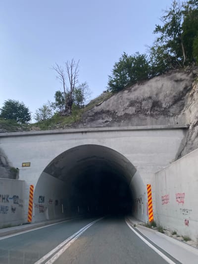 The image shows a road leading into a concrete tunnel, located in the city of Šavnik, Montenegro. The tunnel is illuminated by interior lights, casting a dark shadow within. The exterior of the tunnel is adorned with graffiti, and caution signs are posted on either side. The road itself is paved and marked with white lines. The surrounding landscape features a rocky hillside covered with trees. The sky is clear and pale blue.