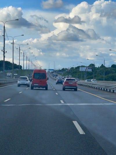 A vertical shot shows traffic on a multi-lane highway. The sky with blue and white clouds is on the left. Rows of street lights line the left side of the road. There are several cars, including a red minivan, a white SUV, and a red sedan, driving in the same direction. The highway appears to be wet. Vegetation can be seen along the edge of the highway. Based on the location, the scene depicts a highway near Strelkovo, Russia.