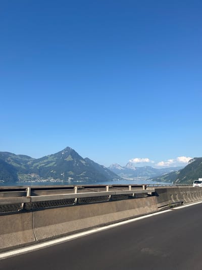 The image displays a daytime scene from a highway overlooking Lake Lucerne (Vierwaldstättersee) in Beckenried, Switzerland. In the immediate foreground, a dark asphalt road surface with a continuous white lane marking on its right edge occupies the lower quarter of the frame. Immediately adjacent to the road is a multi-layered concrete and metal highway barrier. The barrier consists of a concrete base topped by a steel guardrail system with horizontal beams and a mesh infill, extending horizontally across the lower third of the image.
Beyond the barrier, the calm waters of Lake Lucerne stretch across the mid-ground. On the opposite side of the lake, densely forested mountains rise, featuring lighter green cultivated sections on their slopes, with residential structures of villages or towns visible along the shoreline at their base. In the distant background, additional mountain ranges with rugged, rocky peaks and sparse cloud cover are visible under a clear, bright blue sky that dominates the upper half of the image. A white vehicle, possibly a van, is partially visible on the far right, positioned on an unseen road or lane seemingly parallel to the observer's location. No people are visible.