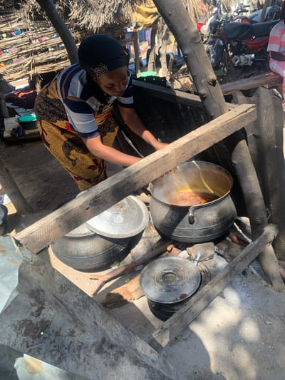 A woman, wearing a headscarf and a patterned dress, is tending to a pot of stew cooking over an open fire. The pot is large and black, and steam is rising from it. The woman is reaching towards the pot with her left hand.
The scene appears to be outdoors, possibly in a village or rural area, given the rustic construction of the cooking area with wooden beams and a dirt floor. There are other metal pots and lids scattered around, as well as some debris. In the background, a pile of motorcycles and other items suggest a busy or cluttered environment. The lighting suggests it is daytime, with bright sunlight casting shadows.
This image captures a moment of everyday life, highlighting the preparation of food in a traditional setting. The woman's posture and the presence of the cooking pot indicate an activity of sustenance. The overall impression is one of resourcefulness and resilience in a simple setting. There is no visible text in the image.
