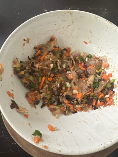 A white plate containing remnants of a chopped vegetable mixture sits off-center on a brown surface. The mixture prominently features diced carrots, green onions, and what appears to be a translucent, gelatinous substance, possibly shredded daikon radish or some kind of noodle. Scattered pieces are visible around the main pile, adding to the sense of being leftovers. The plate is lightly stained with brown spots.
