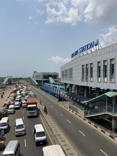 The image presents an elevated view of Alaba Station in Alaiyabiagba, Nigeria, under a bright, partly cloudy sky. The prominent Alaba Station building, a multi-level modern grey structure with "ALABA STATION" in large blue lettering on its roof, is situated on the right, featuring numerous rectangular windows and an elevated platform. A blue and white multi-car train is visible stationary on the elevated tracks at the station. Elevated pedestrian walkways and bridges, one with a green canopy, connect to the station and span across the primary road. Below, a multi-lane highway or major road extends through the scene. The left side of the road exhibits heavy traffic congestion, comprising numerous white vans, cars, motorcycles, and larger trucks, many appearing stationary. Alongside this congested section, multiple market stalls with various colored umbrellas (green, white, striped) are set up. Numerous individuals are seen walking among the vehicles and market stalls, indicative of commercial activity. On the right side of the main road, adjacent to the station, traffic is lighter, with a few groups of people standing on the sidewalk near a stairwell leading to a pedestrian bridge. At least 20 individuals are identifiable across the scene, engaged in transit and market-related movements.