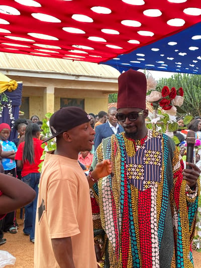 The image appears to be taken at an outdoor event in Kuru Dungus, Nigeria. A man in a colorful, patterned robe and a maroon hat is speaking into a microphone. He wears sunglasses and stands amidst floral decorations. A second man, wearing a black baseball cap and a light-colored shirt, looks up at the speaker. An audience is visible in the background. A red and blue decorative hanging with white circles is on the left side of the image. A building can be seen in the background. Attendees are seated in the foreground.