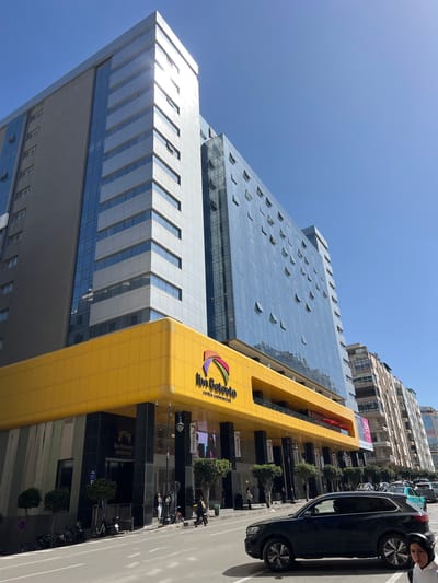The image displays the Ibn Batouta Centre Commercial, a multi-story modern building located in Tanger, Morocco, under a clear blue sky. The building features a prominent, rounded yellow facade on its lower levels, with "Ibn Batouta centre commercial" and a multi-colored logo visible. Below the yellow section, the ground floor entrance areas are clad in black marble-like panels, with signs indicating "Ibn Batouta Bureaux."
Above the yellow base, the building's upper stories are constructed with a mix of light grey paneling and extensive blue-tinted glass curtain walls. Numerous windows on these upper floors are partially open, angled outwards. To the left, another section of the building exhibits darker grey and blue glass components. The sun reflects brightly off the building's surfaces.
On the street level, a wide sidewalk runs alongside a paved road. Several individuals are present on the sidewalk, some congregating near the main entrances of the commercial center, and others walking. A black Volkswagen Touareg SUV is in motion on the road in the foreground, with its driver visible through the window. Other vehicles are visible in the background traffic. Small, manicured trees line sections of the sidewalk, and several motorcycles are parked near the building. Streetlights and banners displaying the Ibn Batouta logo are also present. Signs for businesses such as "Zara" and "Burger King" are visible on the building's exterior. In the background to the right, multi-story residential and commercial buildings with varied architectural styles line the street.