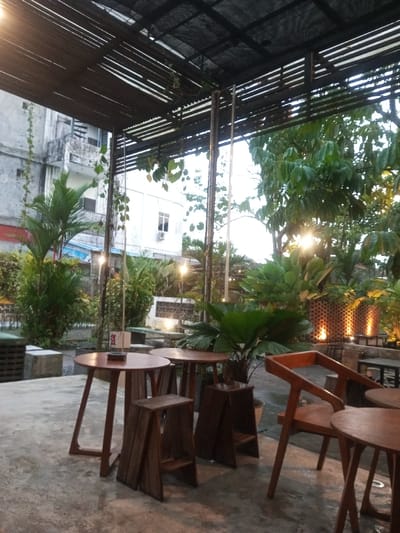 The image displays an unoccupied outdoor cafe or restaurant area in Pontianak, Indonesia, under twilight or overcast conditions. The ground is concrete, leading to various seating arrangements. An overhead structure composed of horizontal wooden slats and a black mesh canopy covers the space, supported by metal poles, some adorned with green vines. In the foreground, two round wooden tables are present, each accompanied by a distinct, geometric wooden stool; one table displays a small, upright menu stand. To the immediate right, a wooden chair with an angled back and a smaller round table are partially visible. The area is heavily landscaped with diverse tropical vegetation, including potted plants and larger trees, illuminated by numerous string lights and other artificial fixtures. In the background, an older, weathered multi-story building with an exterior spiral staircase is visible on the left, adjacent to a lighter-colored building featuring an air conditioning unit. Additional outdoor seating and decorative walls with embedded lighting are discernible deeper within the scene. No individuals or specific actions are depicted.