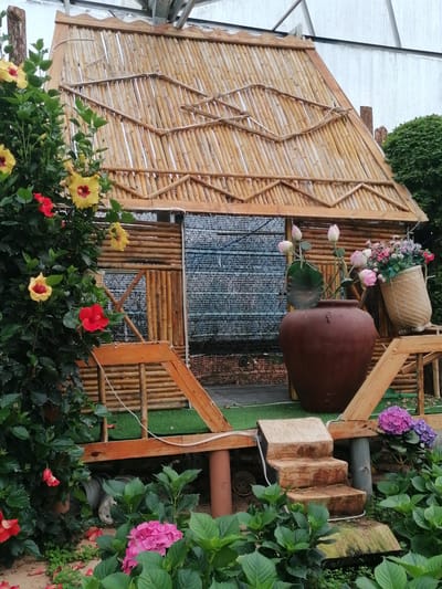 This image presents a serene and vibrantly decorated garden scene, likely set in Lạc Dương, Vietnam, given the cultural context of the architecture and flora. The central subject is a charming, rustic hut or structure, meticulously crafted from bamboo and natural wood. Its steeply pitched roof is a prominent feature, constructed from numerous bamboo poles laid side-by-side, with additional decorative bamboo patterns on its surface. The front of the hut also incorporates horizontal bamboo logs, partially covered by a dark, mesh-like screen or curtain, suggesting an enclosed space.
The hut stands on a raised wooden platform covered with artificial green turf, and three rustic steps, carved from solid logs, lead up to it from the ground. A simple wooden railing surrounds the front of the platform.
The scene is rich with lush plant life. On the left, a large, leafy bush is adorned with numerous hibiscus flowers in striking shades of bright yellow with deep red centers, and vibrant red. Towards the bottom of the frame, more red petals are scattered on the ground, and dense green foliage is visible, including vibrant pink and purple hydrangea bushes blooming below the hut's platform.
To the right of the hut's entrance, a large, rounded brown ceramic pot holds a beautiful arrangement of pink lotus buds, complete with their distinct green leaves and seed pods, adding an element of calm beauty. Next to this pot, a woven bamboo basket overflows with a colorful assortment of artificial flowers, predominantly in shades of pink, purple, and white.
The lighting suggests it is daytime, possibly overcast, contributing to the soft and even illumination of the scene. In the background, hints of a larger structure with metal beams are visible against a light sky, possibly indicating a greenhouse or a park setting. There are no people visible, and no discernible text appears in the image. The entire composition exudes a peaceful, natural, and inviting atmosphere.