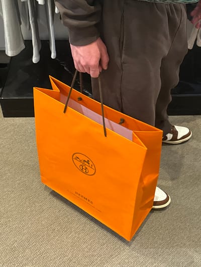 The image depicts an indoor scene in Winchester, United States of America, showing the lower half of a person holding a shopping bag. The individual wears a dark brown long-sleeved top and matching dark brown sweatpants or joggers, paired with white sneakers featuring brown accents. The person's right hand holds a bright orange Hermès shopping bag by its dark brown rope handles. The bag prominently displays the Hermès horse-drawn carriage logo with an "H" and the brand name. Light pink or white tissue paper is visible within the bag. The floor is covered with a light gray textured carpet. In the background, a black, reflective display surface is partially visible, along with blurred light-colored clothing items on a rack to the left.