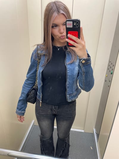 A young Caucasian woman with blonde hair is taking a mirror selfie in an elevator. She is wearing a denim jacket, a black t-shirt, and dark wash flared jeans. She has a small black quilted purse with a gold chain strap slung over her shoulder. Her phone, which has a red and black case, is held up to take the picture. The elevator has beige walls and a metal railing. The floor is grey with a speckled pattern. The location is an elevator in Murino, Russia.