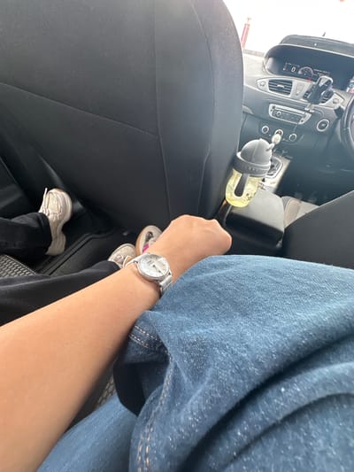 The image depicts two individuals seated in the interior of a car, likely in the city of Trafford, United Kingdom. The foreground is dominated by the left arm of one person, adorned with a white-faced wristwatch featuring a silver band. This arm rests on blue denim jeans. To the left of this individual's arm are the feet of another person, wearing white sneakers and dark trousers. The background reveals the car's dashboard, including a digital display, air vents, and a car's gear shift. A child's sippy cup with a yellow liquid is visible in a cup holder. The overall scene suggests a candid moment captured while traveling in a vehicle.