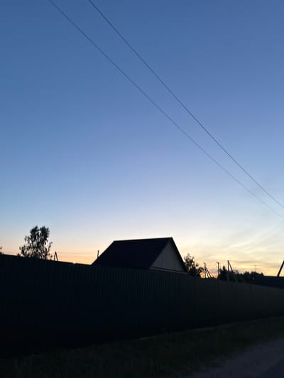 The image, when correctly oriented, presents a vast sky scene at either sunrise or sunset over a rural or semi-rural landscape. The sky dominates the frame, displaying a gradient from a clear, deep blue in the upper and left portions to a lighter, yellowish-orange tone near the horizon on the right. Two thin, dark power lines stretch diagonally across the upper-left sky.
The foreground elements are rendered in silhouette, indicative of the low sun position. A dark, corrugated fence runs horizontally across the lower-right side of the image, bordering a dark strip of ground or low vegetation. Behind the fence, a silhouetted building, possibly a house or shed with a distinct pitched roof, is partially visible on the right. Scattered along the horizon, dark silhouettes of trees and bushes are visible, with some branches extending above the fence and building. In the far distance on the right, several faint, dark shapes suggest utility poles or other structures against the lighter sky.
The scene captures the peaceful transition of day, with no human activity or individuals visible. The weather appears clear. No visible text is present in the image. The setting suggests an open area with simple structures, aligning with the provided location context of Лабжанскі сельскі Савет, Belarus.