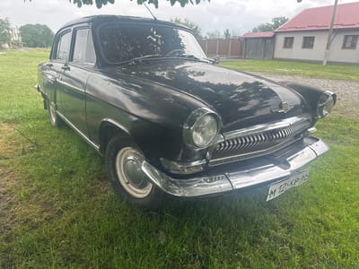 A vintage, black Volga car is parked on a grassy area in front of a house with a red roof. The car is facing the right, with its front prominently displayed. The license plate is visible and reads "M 125KP 65". The house in the background appears to be a residential building. The sky is overcast, suggesting cloudy weather. The scene is set in the city of Dzuarikau, Russia.
