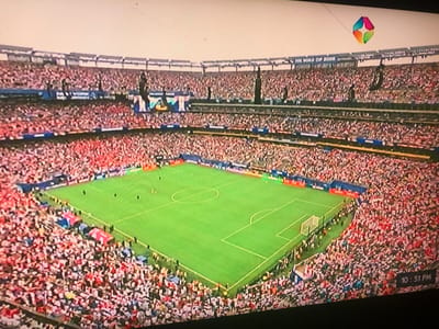 An aerial, wide-angle shot captures a bustling soccer stadium filled with a vast crowd. The bright green soccer field is visible in the center, with white markings and a goalpost. Spectators, dressed in an array of colors, fill the tiered seating that surrounds the field. Some areas of the stands are densely packed, while others appear slightly less so. In the upper right corner, a colorful, diamond-shaped logo hangs in the sky, tethered by a thin line. The sky is overcast and pale. The bottom right corner of the image displays a timestamp: "10:51 PM". There are no discernible details that connect this scene to Rukuba Road, Nigeria.