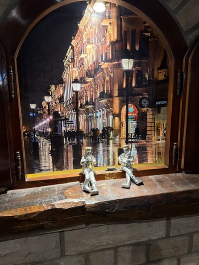 The image shows a view through an arched wooden window frame onto a nighttime city street scene. The street is wet, reflecting the lights from buildings and streetlamps. People are visible walking along the sidewalk, some with umbrellas. Two small, silver-colored figurines are seated on the wooden windowsill in the foreground. One figurine is playing a trumpet, and the other is holding a drum. The background depicts buildings with ornate architecture, illuminated by numerous lights. The scene is set in the city of Kumbor, Montenegro, as indicated by the presence of "Perspective" signage on one of the storefronts, which is a known establishment in Kumbor.