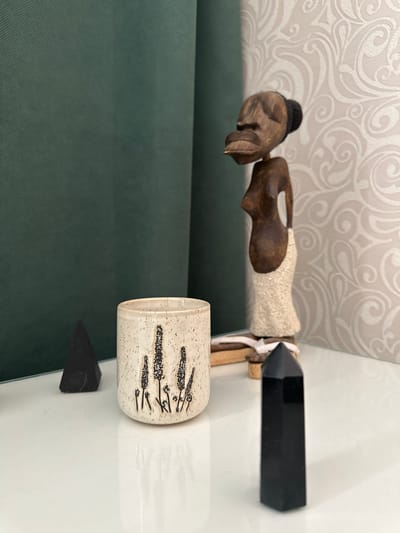 A still life scene on a white, reflective surface features several decorative objects. In the foreground, a tall, black, faceted crystal obelisk stands to the right. To the left of it, a small, dark, four-sided pyramid-shaped object is visible. Between these two crystals sits a short, speckled ceramic cup adorned with black illustrations of tall, slender plants. Behind the cup, a wooden figurine of a woman with pronounced facial features and wearing a textured skirt stands prominently. The background consists of a dark green curtain on the left and a wallpaper with an ornate white scroll pattern on the right. The overall setting appears to be indoors, possibly a home or decorative display area. There is no information or indication that this scene is located in Malye Ust'ya, Russia.