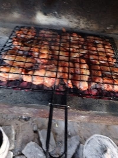 In Kasri Arifan, Uzbekistan, the scene depicts a close-up, downward-angled view of meat being grilled. A rectangular black metal wire grill is densely loaded with numerous pieces of light reddish-brown meat, consistent with poultry cuts, cooking directly above glowing red charcoal briquettes. A black metal handle extends from the center of the grill towards the bottom of the frame. The grilling process is taking place within a structure composed of rough, grey stones or bricks visible in the lower foreground. The upper background shows a dark, possibly soot-stained wall or interior surface. A light-colored object is partially visible on the far-left foreground, and a circular, possibly wire-mesh object is partially visible on the far-right foreground. No individuals are present.